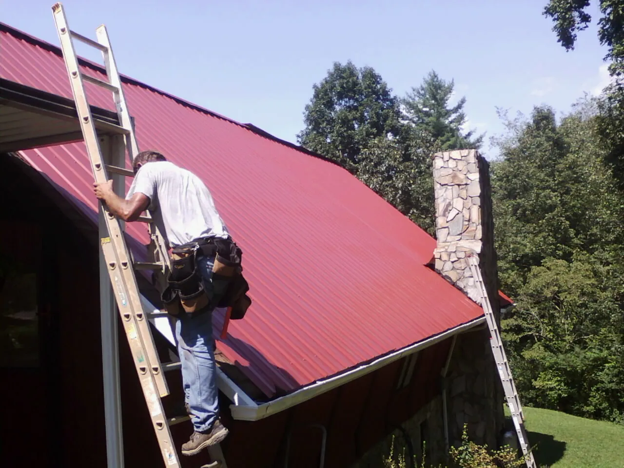 Roofer on a ladder beside a red metal roof and stone chimney — Soffit & Fascia Repair by Roofing Brothers in Bourbonnais